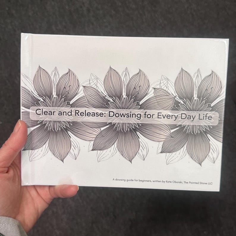 May include: A white book with the title "Clear and Release: Dowsing for Every Day Life" is held in a hand. The cover features three large, grey floral illustrations. Below the title is the text "A dowsing guide for beginners, written by Kate Oborski, The Pointed Stone LLC."