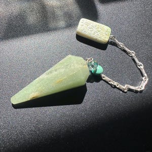 May include: A light green, cone-shaped gemstone pendulum attached to a silver-toned metal chain. The chain features a small, round turquoise bead and a clear glass bead, ending in a rectangular, light green stone fob with a carved floral pattern.
