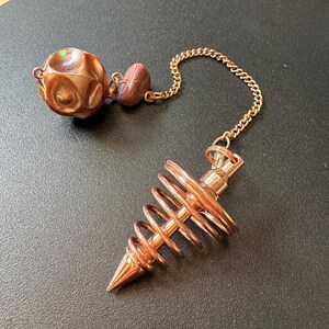 May include: A copper-toned metal pendulum featuring a spiral cone-shaped weight attached to a chain. The chain connects to a smooth, round, iridescent copper-colored bead and a smaller, matte, mauve-colored stone bead at the end of the chain.