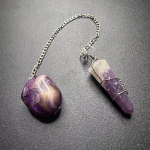 May include: A pendulum featuring a silver-toned metal chain connected to a smooth, rounded purple and gold marbled stone bead on one end and a wire-wrapped, pointed amethyst crystal pendant on the other end, showcasing natural purple and white hues.