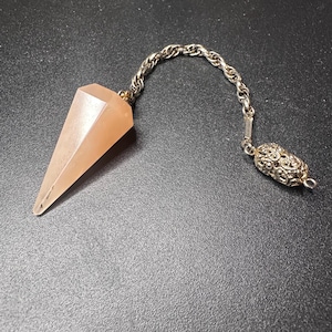 May include: A faceted, light peach-colored stone pendulum attached to a silver-toned metal chain. The chain features a twisted link design and terminates in an ornate, filigree-style metal bead at the end, providing a decorative finish.