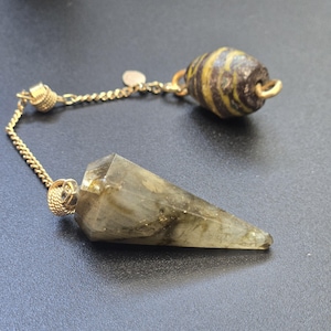 Puede incluir: Un péndulo con una punta de piedra gris verdosa translúcida y facetada unida a una cadena de metal dorado. La cadena se conecta a una pequeña cuenta marrón oscuro con rayas horizontales amarillas y un diminuto dije de metal plano en forma de corazón.