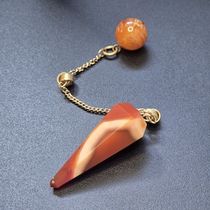 Puede incluir: Un péndulo de radiestesia con una punta de piedra facetada en forma de cono con bandas naranjas y blancas, unida a una cadena de metal dorado que termina en una pequeña cuenta de piedra naranja pulida y redonda en el otro extremo.