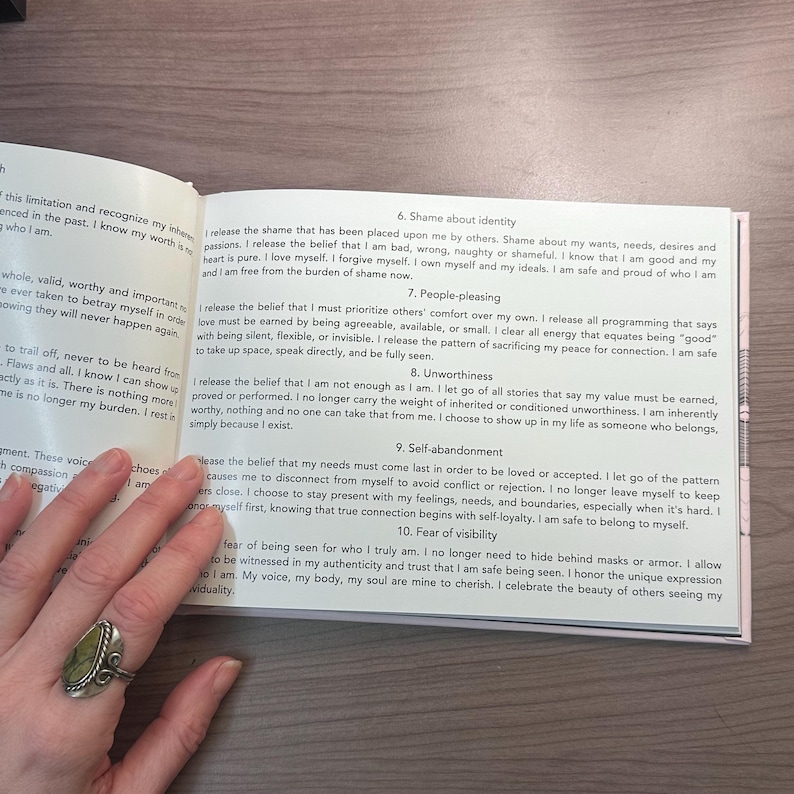 May include: An open book with text on a pink page, discussing self-love and releasing shame. A hand with a ring is holding the book. The text is in English.