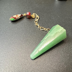 Könnte beinhalten: Ein grüner, facettierter, kegelförmiger Steinanhänger an einer goldfarbenen Metallkette. Die Kette ist mit einer kleinen, dunkelroten, runden Perle und einem tropfenförmigen Anhänger mit roten, weißen und grünen Emaille-Blumenmustern versehen.