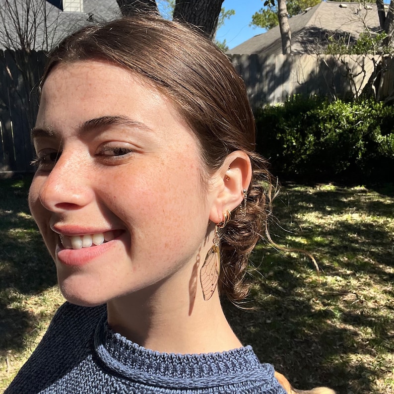 May include: A close-up shot of a person wearing gold hoop earrings and delicate, dangling earrings with a wing-like design. The earrings are a light, translucent color. The person is wearing a blue sweater.