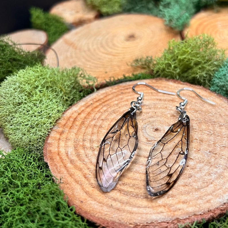 May include: A pair of silver-toned dangle earrings featuring delicate, translucent wing-shaped pendants. The wings have intricate vein detailing and are set against a backdrop of wood slices and green moss.