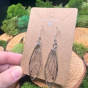 May include: A pair of handmade earrings featuring clear, wing-shaped pendants with intricate black vein detailing, suspended from silver-colored hooks. The earrings are displayed on a brown card with the word "Handmade" and a heart symbol. The background includes green moss and wood slices.