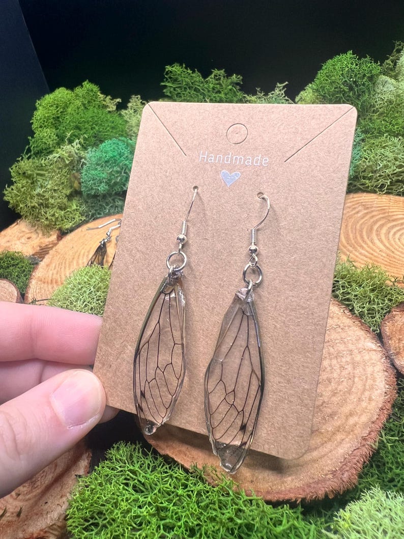 May include: A pair of handmade earrings featuring clear, wing-shaped pendants with intricate black vein detailing, suspended from silver-colored hooks. The earrings are displayed on a brown card with the word "Handmade" and a heart symbol. The background includes green moss and wood slices.