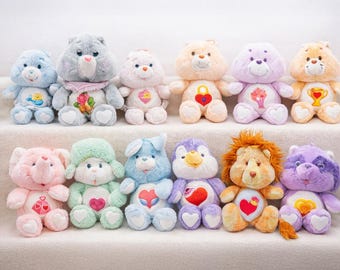 May include: A collection of plush Care Bears in various colors, including blue, gray, pink, peach, purple, and orange. Each bear has a unique belly badge and is arranged on a white surface.
