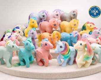 May include: A collection of vintage My Little Pony plastic toy figures in various pastel colors including pink, blue, yellow, and white. Each pony features a unique molded mane, tail, and symbol on its flank. A circular blue logo reads The Local 11.