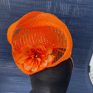 May include: Orange mesh hat with a large orange flower accent. The hat is on a black hat form.