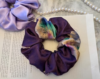 May include: A light lavender satin scrunchie and a dark purple satin scrunchie with a multi-colored floral and abstract pattern. The dark purple scrunchie features a subtle embossed texture throughout the fabric, creating a gathered, circular shape.