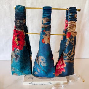 May include: Three fabric headbands featuring floral patterns in shades of teal, red, and cream. Each headband has a wide front section and a gathered elasticized back strap, displayed on a gold-toned metal stand with a marble-patterned base.