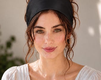 May include: A wide, solid black fabric headband worn across the forehead. The headband is made of a smooth, matte material and features a simple, unadorned design that sits securely against the hair, providing a clean and functional hair accessory.