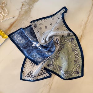 May include: A square silk scarf featuring a multi-colored paisley pattern in shades of light blue, sage green, and white. The scarf has a dark navy blue border and is decorated with intricate floral motifs and geometric shapes throughout the fabric.