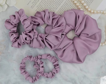 May include: Four purple satin scrunchies, two large and two small, on a marble surface. The scrunchies are made of soft, silky fabric and are perfect for holding hair in place.