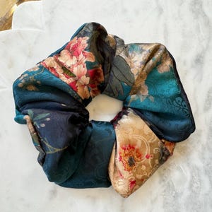 May include: A circular hair scrunchie featuring a multi-colored floral pattern on a dark teal and gold background. The fabric displays a satin-like sheen with intricate botanical illustrations, including pink and red flowers, dark green leaves, and swirls.