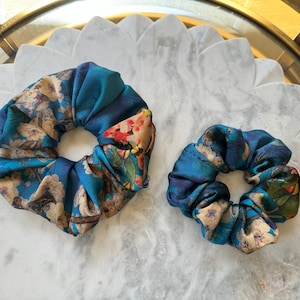 May include: Two blue and beige patterned scrunchies. The larger scrunchie has a floral pattern with red, yellow, and green accents. The smaller scrunchie has a similar pattern but is smaller in size.