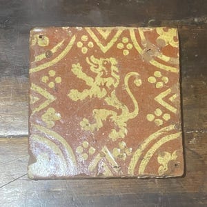 May include: Square, antique tile with a red background and yellow design. The design includes a heraldic lion, floral elements, and geometric shapes. The tile has a worn, aged appearance, with small holes in the corners.