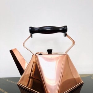 May include: A polished copper tea kettle with a distinct geometric, pyramid-like shape. It features a black handle with a curved grip and a small black knob on the lid. The spout is angled and extends from the side of the metallic body.