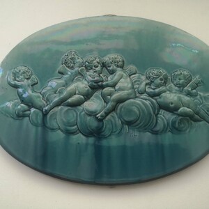 May include: Oval, teal ceramic plaque with a relief sculpture of cherubs on clouds. The cherubs are in different poses, some holding objects. The plaque has a glossy finish and a subtle iridescent sheen.