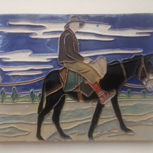 May include: A ceramic tile depicting a person riding a donkey. The rider wears a hat, jacket, and pants, with red socks. The donkey is black, and the background features a blue sky with white clouds and a landscape.