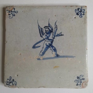 Puede incluir: Azulejo cerámico cuadrado con un diseño pintado a mano de un querubín alado sosteniendo una antorcha, en azul sobre fondo blanco. Motivos florales decorativos en cada esquina. El azulejo tiene un aspecto envejecido y desgastado.