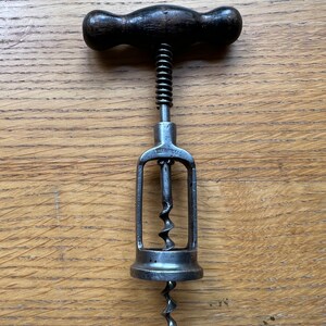 May include: A vintage corkscrew with a dark wooden handle and a metal frame. The corkscrew features a spiral worm and a spring mechanism. The handle is a dark brown colour, and the metal components have a silver finish. It is designed for removing corks from wine bottles.