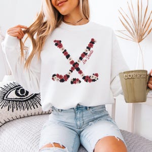 May include: White sweatshirt featuring two crossed hockey sticks made of burgundy, white, and black flowers. The words "Go Devils" are written in burgundy script. The sweatshirt is a casual, comfortable style.