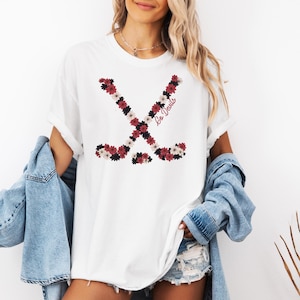 May include: White t-shirt featuring a floral hockey stick design in red, black, and white. The words "Go Devils" are printed in a script font on the shirt. The shirt is worn with a denim jacket.