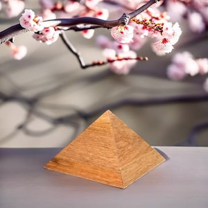 Pode incluir: Uma pirâmide de madeira com acabamento marrom claro está sobre uma superfície cinza. A pirâmide está em foco, enquanto o fundo está desfocado e apresenta um ramo de flores cor-de-rosa.