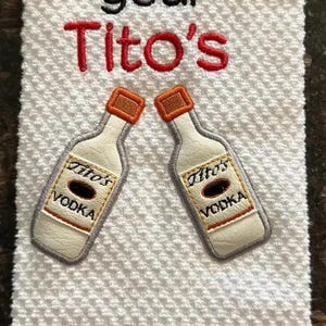 May include: White kitchen towel with gray and white stripes. The towel has embroidered text that says "Show me your Tito's" with two embroidered images of Tito's Vodka bottles.