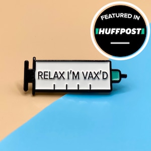 Könnte beinhalten: Eine schwarze und weiße Emaille-Anstecknadel in Form einer Spritze mit dem Text "Relax I'm Vax'd" in schwarzen Buchstaben. Die Anstecknadel befindet sich auf einem hellbraunen und blauen Hintergrund.