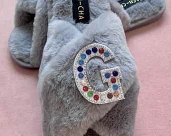 May include: Gray fuzzy slippers with a large, sparkly, multi-colored letter G on the front. The slippers have a criss-cross strap design and the brand name "Bach-Cha" is printed on the side.