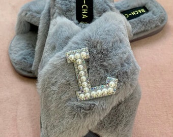 May include: Gray fuzzy slippers with a pearl-embellished letter L on the front. The slippers have a criss-cross strap design and say "BACH-CHA" on the side.