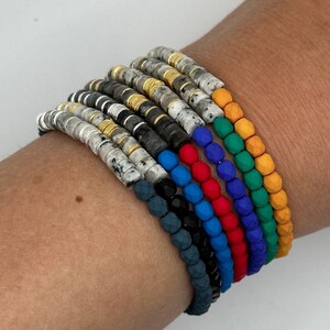 Può includere: Un set di otto braccialetti colorati con perline. I braccialetti sono impilati sul polso di una persona. I braccialetti sono realizzati con diversi materiali, tra cui perline, pietre e metallo. I braccialetti sono di vari colori, tra cui nero, blu, rosso, verde e giallo.