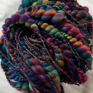 May include: A skein of hand-spun art yarn featuring a thick and thin texture with a vibrant multi-colored palette. The yarn displays blended hues of deep purple, magenta, teal, emerald green, and golden yellow, twisted into a complex, irregular ply.