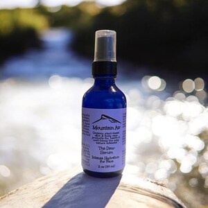Pu&ograve; includere: Una bottiglia di vetro blu di "The Dew Serum" di Mountain Air, con un tappo spray nero. L'etichetta include il testo "Intense Hydration for Face" e la bottiglia contiene 59 ml di prodotto.