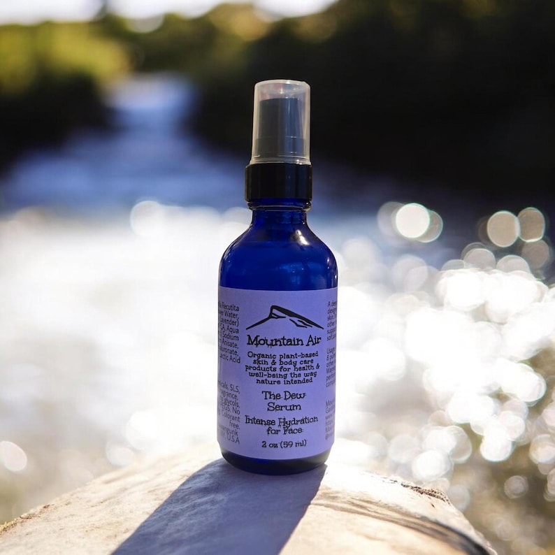 Pu&ograve; includere: Una bottiglia di vetro blu di "The Dew Serum" di Mountain Air, con un tappo spray nero. L'etichetta include il testo "Intense Hydration for Face" e la bottiglia contiene 59 ml di prodotto.