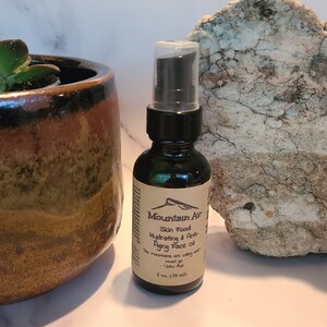 May include: A dark glass bottle of Mountain Air Skin Food Hydrating & Anti-Aging Face Oil, 1 fl oz (30 ml). The label features a mountain graphic. A succulent plant in a brown ceramic pot and a large rock are in the background.