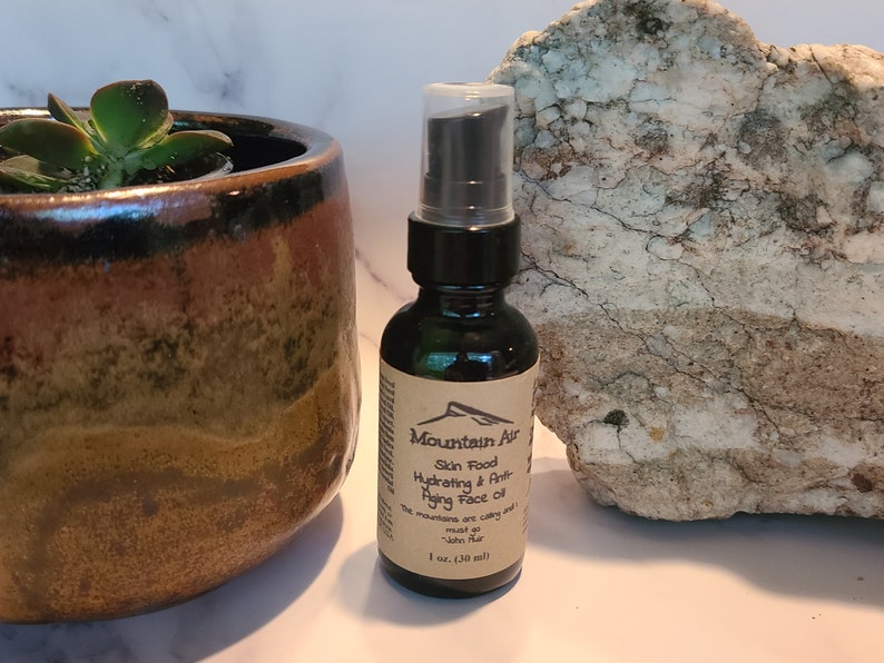 May include: A dark glass bottle of Mountain Air Skin Food Hydrating & Anti-Aging Face Oil, 1 fl oz (30 ml). The label features a mountain graphic. A succulent plant in a brown ceramic pot and a large rock are in the background.