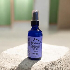Pu&ograve; includere: Una bottiglia di vetro blu cobalto con un nebulizzatore nero. L'etichetta recita "Mountain Air The Dew Serum Intense Hydration for Face". La bottiglia contiene 59 ml di prodotto. Lo sfondo &egrave; sfocato, verde e bianco.