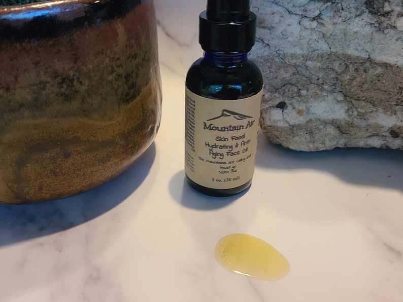 May include: A dark blue glass bottle of Mountain Air Skin Food Hydrating & Anti-Aging Face Oil, with a brown label. A small amount of oil is spilled on the white surface. The bottle contains 1 fl oz (30 ml).