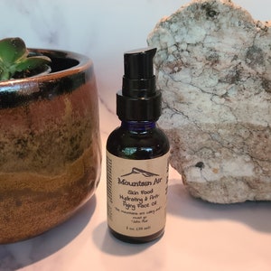 May include: A dark blue glass bottle with a black pump dispenser, labeled "Mountain Air Skin Food Hydrating & Anti-Aging Face Oil." The bottle is 1 oz (30 ml). A succulent plant in a brown ceramic pot and a white and gray rock are in the background.
