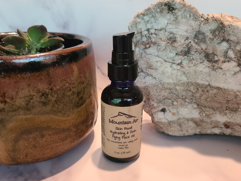 May include: A dark blue glass bottle with a black pump dispenser, labeled "Mountain Air Skin Food Hydrating & Anti-Aging Face Oil." The bottle is 1 oz (30 ml). A succulent plant in a brown ceramic pot and a white and gray rock are in the background.