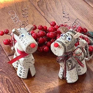 May include: Two handmade reindeer figurines crafted from wine corks, featuring googly eyes, red noses, and decorative ribbon. The reindeer have wire antlers and are positioned near a cluster of red berries, perfect for holiday decor.