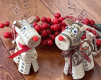 May include: Two handmade reindeer figurines crafted from wine corks, featuring googly eyes, red noses, and decorative ribbon. The reindeer have wire antlers and are positioned near a cluster of red berries, perfect for holiday decor.