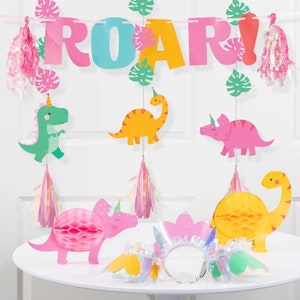 Op de afbeelding: Een roze, gele en groene dinosaurus-partybanner met het woord "ROAR!" in kleurrijke letters. De banner is versierd met groene bladeren en roze en gele kwastjes. Er zijn ook verschillende dinosaurus-uitknipsels, waaronder een groene dinosaurus, een gele dinosaurus en een roze dinosaurus. De dinosaurussen dragen allemaal feestmutsen.