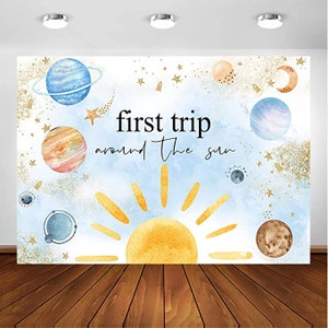 May include: A blue and white watercolor backdrop with a yellow sun and planets. The text "first trip around the sun" is written in black. This backdrop is perfect for a baby shower or birthday party.